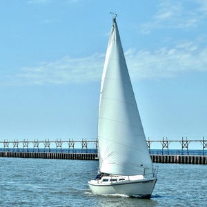 May include: A white sailboat with a single sail is sailing on a calm blue body of water. The sailboat is in front of a pier with a wooden railing.