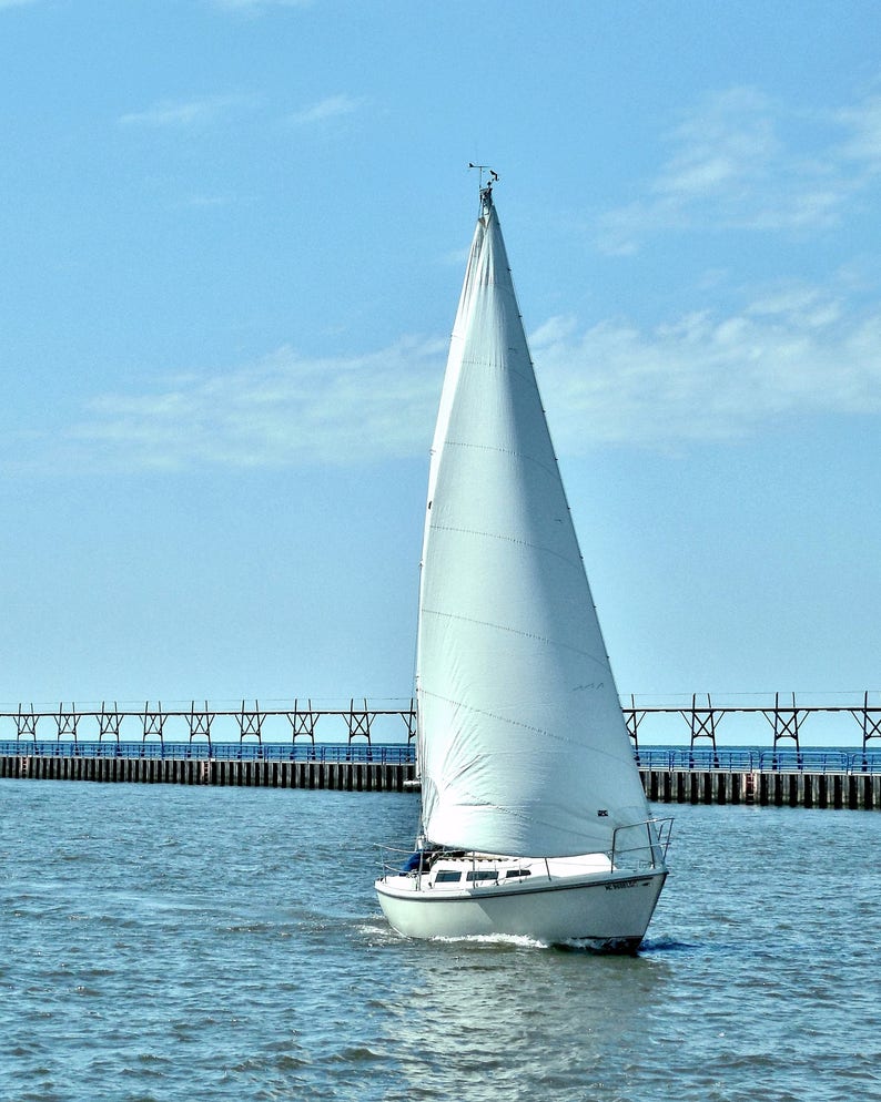 May include: A white sailboat with a single sail is sailing on a calm blue body of water. The sailboat is in front of a pier with a wooden railing.