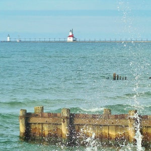 May include: A red and white lighthouse stands on a pier extending into a blue body of water. Waves crash against a wooden breakwater in the foreground.