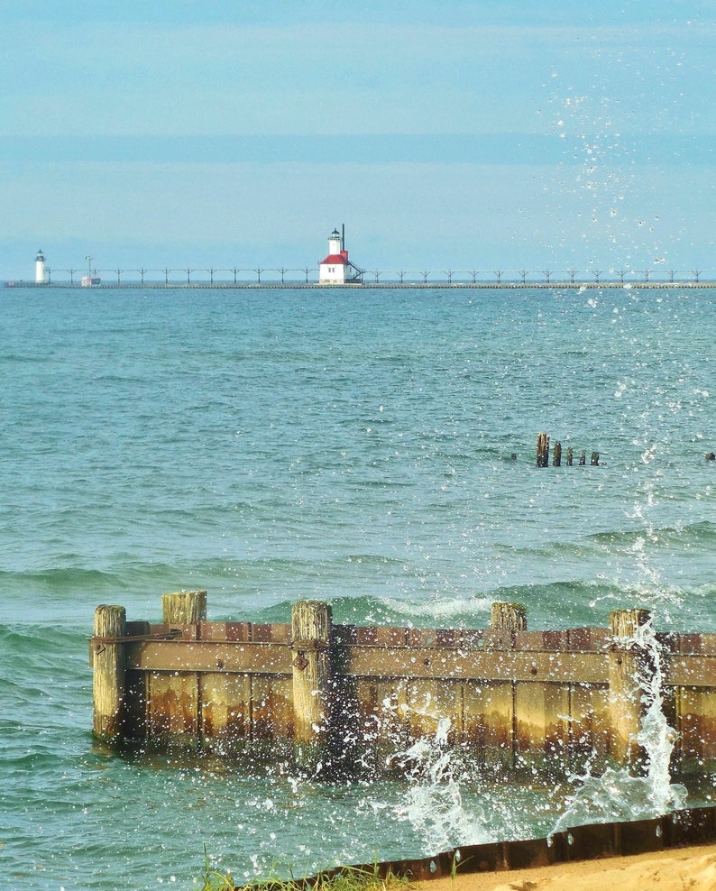 May include: A red and white lighthouse stands on a pier extending into a blue body of water. Waves crash against a wooden breakwater in the foreground.