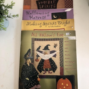 May include: A stack of Halloween-themed books and patterns. The top book reads "Humble Spirits" with a bird illustration. Other titles include "Hallowing Harvest" and "Making Spirits Bright." The bottom book features a witch doll and a pumpkin pillow.