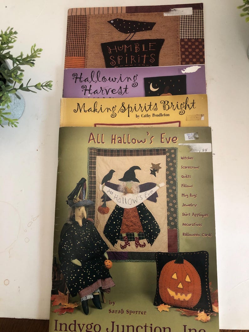 May include: A stack of Halloween-themed books and patterns. The top book reads "Humble Spirits" with a bird illustration. Other titles include "Hallowing Harvest" and "Making Spirits Bright." The bottom book features a witch doll and a pumpkin pillow.