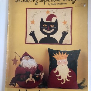 May include: A book cover titled "Making Spirits Bright" by Cathy Pendleton. The cover features a black cat wearing a Santa hat, a Santa Claus figure, and a pillow with a king figure. The text "Indygo Junction, Inc." is at the bottom.