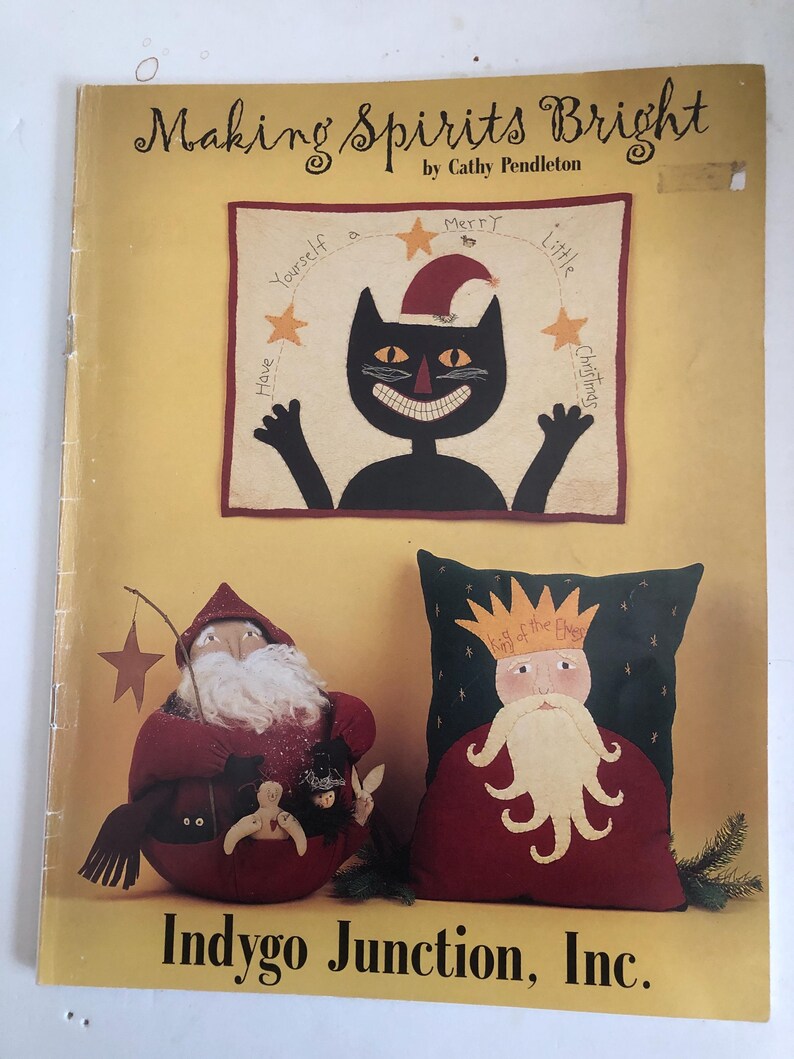 May include: A book cover titled "Making Spirits Bright" by Cathy Pendleton. The cover features a black cat wearing a Santa hat, a Santa Claus figure, and a pillow with a king figure. The text "Indygo Junction, Inc." is at the bottom.