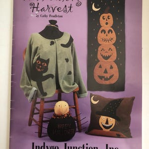 May include: A purple book cover with the title "Hallowing Harvest" and a green jacket with a black cat applique. Other items include a pumpkin banner, a witch pillow, and a spider-like figure with "Happy Halloween" text.