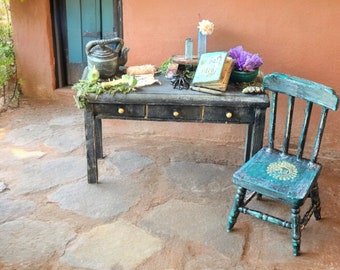 May include: A rustic black desk with three drawers and gold knobs, adorned with a teapot, scrolls, and a book. A teal chair with a sun design sits beside the desk. The scene is set outdoors with a stone floor and a teal door.