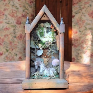 May include: A wooden clock tower with a distressed paint finish. The clock face is surrounded by moss, leaves, and a small unicorn figurine. The tower is sitting on a wooden table.