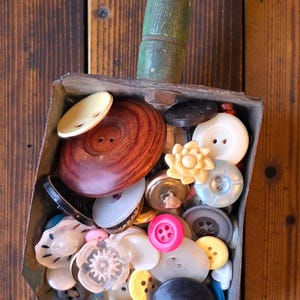 May include: A collection of various buttons in a small, rectangular metal scoop with a green wooden handle. The buttons are in different sizes, colors, and materials, including wood, plastic, and metal. The background is a wooden surface.