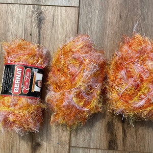 May include: Three balls of Bernat 'Love Cash' yarn in a mix of orange, yellow, and white. The yarn has a fluffy, fuzzy texture.
