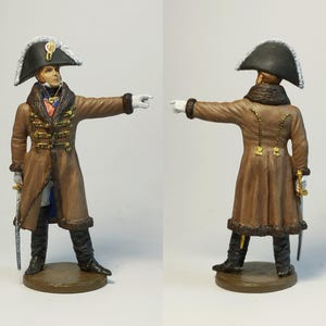 May include: A detailed miniature figurine of a military officer in a brown coat with gold buttons, black boots, and a bicorne hat. The figure is pointing with a white-gloved hand, and a sword is visible at the side. The figure is standing on a circular base.
