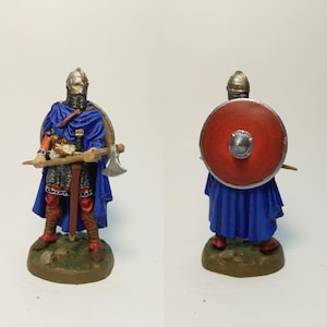 May include: A detailed miniature Viking warrior figure. The figure wears a blue cape, chainmail, and a helmet, and carries an axe and a red shield with a silver boss. The figure is standing on a small, textured base.