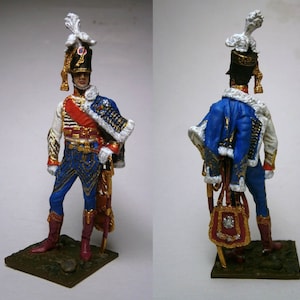 Puede incluir: Figura en miniatura detallada de un oficial militar con uniforme azul con detalles dorados, una faja roja y un sombrero negro alto con plumas blancas. La figura se encuentra sobre una pequeña base verde, mostrando vistas frontal y trasera.