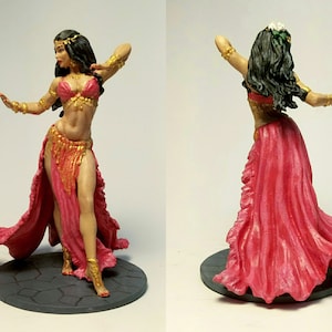Puede incluir: Figura en miniatura de una bailarina con un traje rojo y dorado. La bailarina tiene cabello oscuro y está adornada con joyas doradas. El disfraz incluye un top de sujetador, una falda con abertura y una tela fluida. La figura está sobre una base gris.