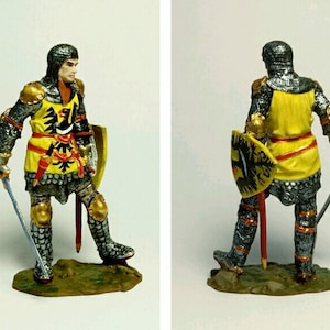 May include: A hand-painted miniature figure of a knight in armour, holding a sword and shield. The knight is wearing a yellow and red tunic with a black eagle on the shield. The figure is standing on a green base.