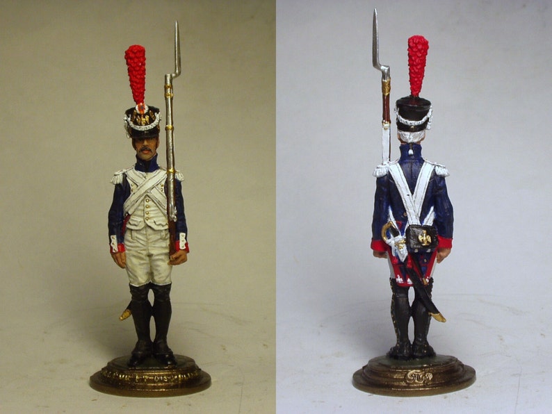 May include: A painted tin soldier figurine wearing a blue and white uniform with a red plume on his hat. He is holding a rifle and standing on a circular base.