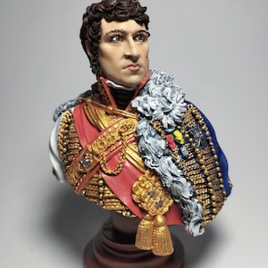 Puede incluir: Busto detallado de un hombre con uniforme militar. La escultura presenta una faja roja, adornos dorados y una capa azul. La figura tiene cabello oscuro y está montada sobre una base marrón. El busto es un objeto decorativo.