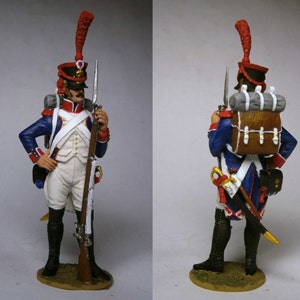 Puede incluir: Una figurita miniatura pintada de un soldado francés de las guerras napoleónicas. El soldado lleva un uniforme azul y blanco con un sombrero rojo y blanco. Lleva un mosquete y una mochila a la espalda.