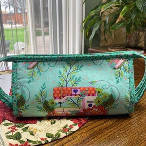 May include: A teal blue fabric zippered pouch with a floral and sewing machine print. The pouch has two fabric handles.