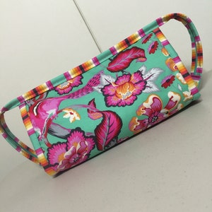May include: A teal fabric tote bag with a floral print in shades of pink, orange, and white. The bag has two handles and a contrasting trim in orange, yellow, and pink.