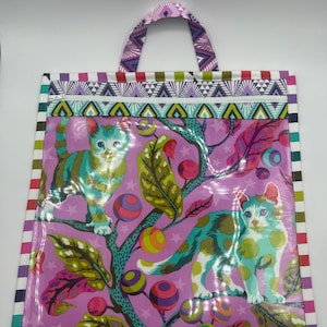 May include: A purple reusable tote bag with a colorful striped border and a handle. The bag features a whimsical design of cats on a branch with colorful orbs and stars.