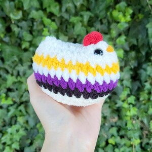 Peut inclure: Un poulet crocheté blanc, jaune, violet, noir et rouge. Le poulet fait à la main a une crête rouge et un œil noir. Le poulet est tenu dans une main sur un fond de feuilles vertes.