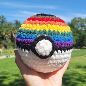 Puede incluir: Una Poké Ball de crochet con un patrón de arcoíris y un centro blanco. La pelota se sostiene en una mano contra un fondo de hierba verde.
