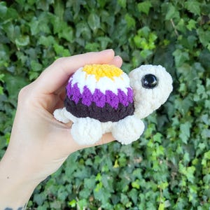 May include: A crocheted turtle toy with a non-binary pride flag design. The shell is yellow, white, purple, and black. The turtle has a cream-coloured body, head, and legs, and black eyes. The background is a green leafy plant.