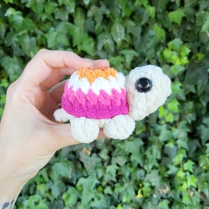 May include: A small, handmade crochet turtle in cream, pink, orange, and white. The turtle's shell features a pink, orange, and white color scheme, while the body and head are cream-colored. The turtle has a black eye.