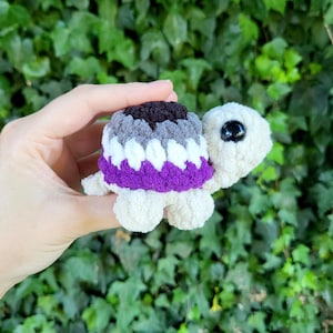 May include: A crocheted turtle with a shell in the colors of the asexual pride flag: black, gray, white, and purple. The turtle has a cream-colored body, four small feet, and a large black eye. The background is a green leafy plant.