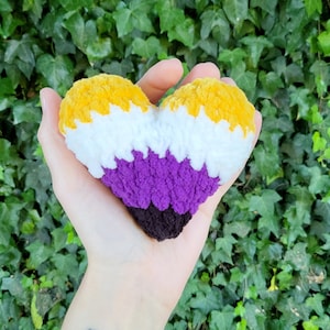 Może przedstawiać: Pluszowa zabawka w kształcie serca wykonana na szydełku w kolorze żółtym, białym, fioletowym i czarnym. Kolory reprezentują flagę dumy osób niebinarnych.