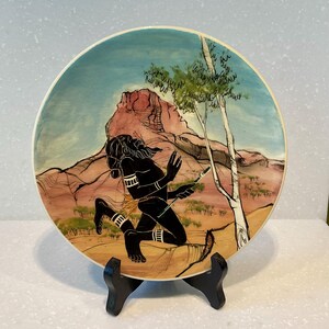 May include: Decorative plate depicting a running black figure in a desert scene. The plate has a painted backdrop of a mountain range and a blue sky. A tree is on the right. The plate is displayed on a black stand. The plate's design is in a circular shape.