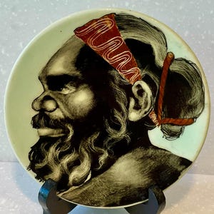 May include: A decorative ceramic plate featuring a profile portrait of a person with a beard and a headband. The portrait is in black and white with a light green border. The headband is red with a white wavy design.