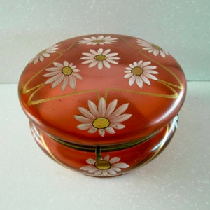 Puede incluir: Una caja de baratijas redonda de color coral con tapa abisagrada. La tapa está decorada con margaritas blancas y detalles dorados. La caja tiene un borde dorado alrededor de la base y la tapa.