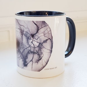May include: White ceramic mug with a black handle and interior. The mug features a watercolor design in shades of gray and black, resembling a cross-section of a tree trunk. The text "LYON ROAD ART" is printed on the side.