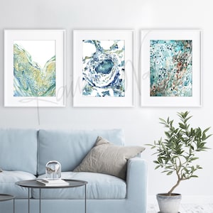 May include: Three abstract watercolor paintings in white frames. The paintings feature blue, green, and gold colors with a swirling, organic pattern.