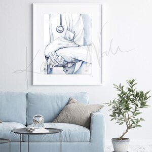 Op de afbeelding: Ingelijste aquarelprint van een arts die de hand van een pati&euml;nt vasthoudt, met een stethoscoop. Het kunstwerk is in blauw- en wittinten. De afbeelding is gemonteerd in een witte lijst en hangt aan een witte muur, wat een rustgevende sfeer cre&euml;ert.