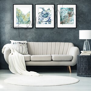 May include: Three framed abstract watercolor paintings in shades of blue, green, and brown. The paintings are hung on a dark gray wall above a white sofa.