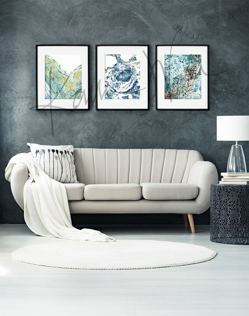 May include: Three framed abstract watercolor paintings in shades of blue, green, and brown. The paintings are hung on a dark gray wall above a white sofa.