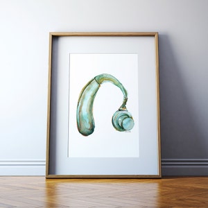 Puede incluir: Una pintura de acuarela de un audífono azul y verde. El audífono se representa de perfil, con el altavoz mirando al espectador. La pintura está enmarcada en un marco de madera claro.
