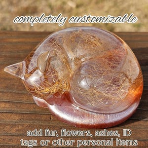 May include: A clear resin cat figurine in a curled sleeping position, containing embedded brown animal fur. The text 'completely customizable' appears at the top, and 'add fur, flowers, ashes, ID tags or other personal items' is at the bottom.