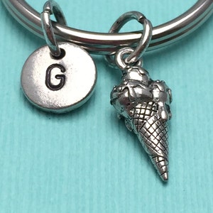 May include: Silver keychain with a letter G charm and a small ice cream cone charm. The keychain is on a turquoise background.