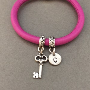 May include: A pink leather bracelet with two charms: a silver key and a silver charm with the letter "C".