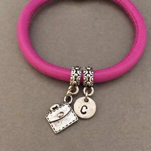 May include: A pink leather bracelet with two silver charms. One charm is a small briefcase and the other is a circle with the letter "C" on it.