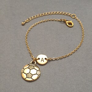 May include: Gold-colored bracelet featuring a soccer ball charm and a small round charm with the letter "A". The bracelet has a delicate chain and an adjustable clasp. The soccer ball charm has black detailing.