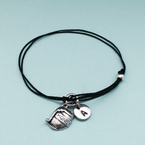 May include: A black string bracelet with a silver honey pot charm and a silver charm with the letter A.