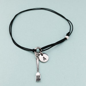 May include: A black string bracelet with a silver fork charm and a silver disc charm with the letter 'A' engraved on it.