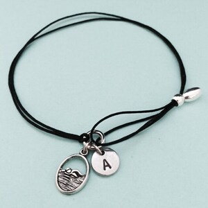 May include: A black adjustable cord bracelet with a silver charm featuring a swimmer and a silver charm with the letter A.