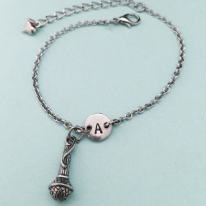 以下が含まれることがあります： 銀色のチェーンブレスレット。銀色のマイクのチャームと、銀色の円盤に「A」の文字が刻まれています。