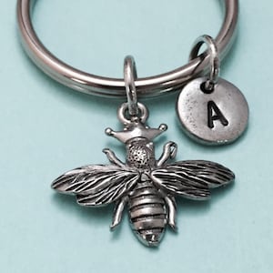 May include: Silver bee keychain with a crown on its head and a silver charm with the letter A. The keychain is on a light blue background.