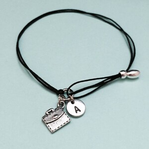 May include: A black string bracelet with a silver charm of a briefcase and a silver charm with the letter A.
